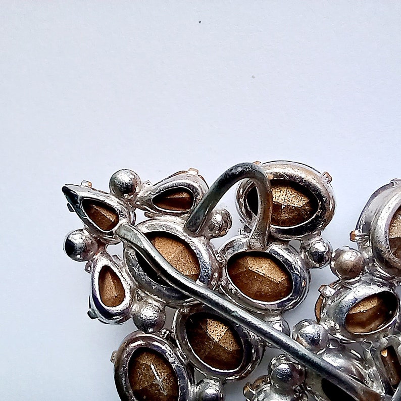 May include: A silver-toned brooch with a cluster of brown and clear rhinestones. The brooch has a decorative pin and a loop for attaching to clothing.