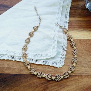 May include: Gold-toned necklace with an ornate, repeating pattern of stylized floral and geometric shapes. The necklace is laid on a white lace-trimmed cloth, set against a wooden surface. The necklace has an adjustable chain.