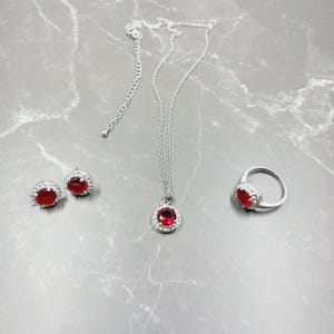 May include: A jewelry set featuring a silver necklace, earrings, and ring. Each piece has a large, round, red gemstone surrounded by smaller clear stones. The jewelry is displayed on a gray marble surface.