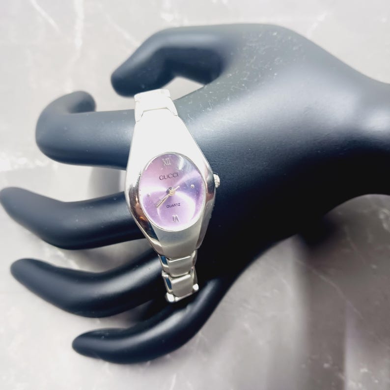 May include: A silver Gucci quartz watch with a purple oval face. The watch has a silver strap and Roman numeral hour markers. The watch is displayed on a black hand-shaped display.