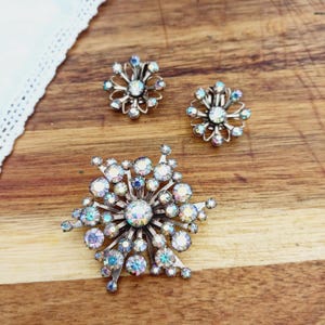 K&ouml;nnte beinhalten: Ein Set aus drei Broschen mit schimmernden Strasssteinen in Blumenform. Die gr&ouml;&szlig;te Brosche ist ein Schneeflockenmuster, die beiden kleineren Broschen sind einfache Blumenmuster.