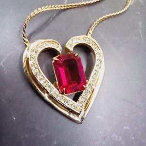 May include: Gold-toned heart pendant necklace with a large, rectangular, ruby-red gemstone at the center. The heart is outlined with small, clear crystals. The chain is a delicate, twisted design.