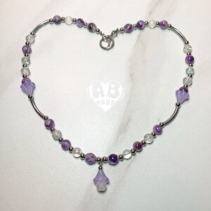 Puede incluir: Un collar morado y plateado con un dije de flor. El collar está hecho con cuentas de amatista y vidrio transparente.