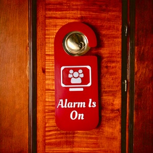 May include: A red door hanger with a white silhouette of people in a monitor and the text "Alarm Is On".