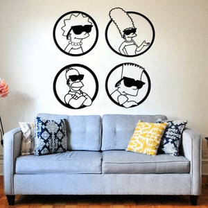 May include: Four black and white silhouette wall decals of the Simpsons family members, Homer, Marge, Bart, and Lisa, wearing sunglasses. The decals are on a white wall above a light gray couch with two pillows. A black tripod floor lamp with a pink flower-shaped shade is in the corner of the room.