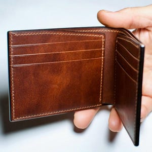 May include: Open brown leather wallet with multiple card slots and a bill compartment. The wallet is held open, showcasing the interior design and stitching details. The leather has a rich, textured appearance.