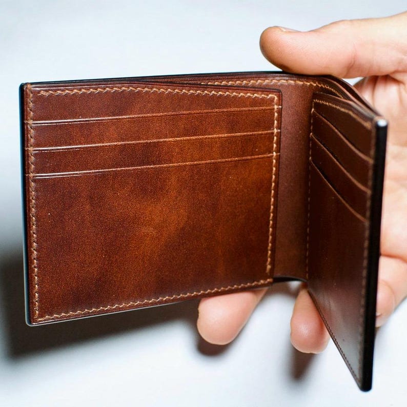 May include: Open brown leather wallet with multiple card slots and a bill compartment. The wallet is held open, showcasing the interior design and stitching details. The leather has a rich, textured appearance.