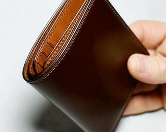 May include: A bifold wallet featuring a smooth, dark brown exterior and a lighter tan interior. The wallet has visible stitching along the edges and multiple interior card slots, showcasing a clean, structured design with a polished finish.