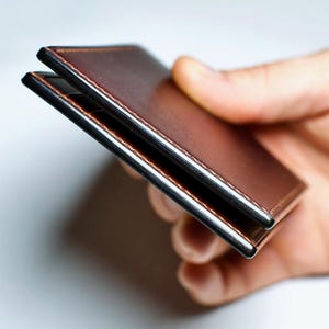 May include: A brown leather wallet with a silver metal frame. The wallet is held in a hand, showcasing its slim profile and stitched edges. The leather has a rich, warm tone.