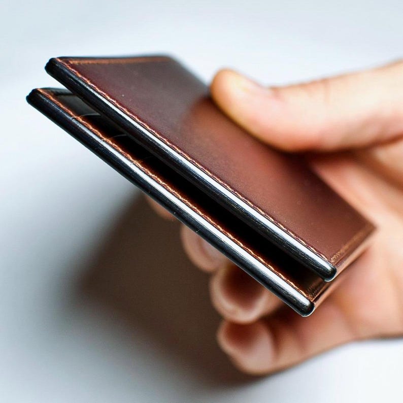 May include: A brown leather wallet with a silver metal frame. The wallet is held in a hand, showcasing its slim profile and stitched edges. The leather has a rich, warm tone.