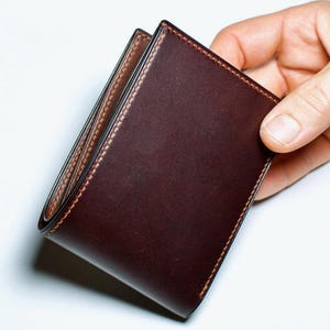 May include: A dark brown leather wallet with a bi-fold design. The wallet has a smooth, polished exterior and contrasting stitching around the edges. The interior is a lighter shade of brown.