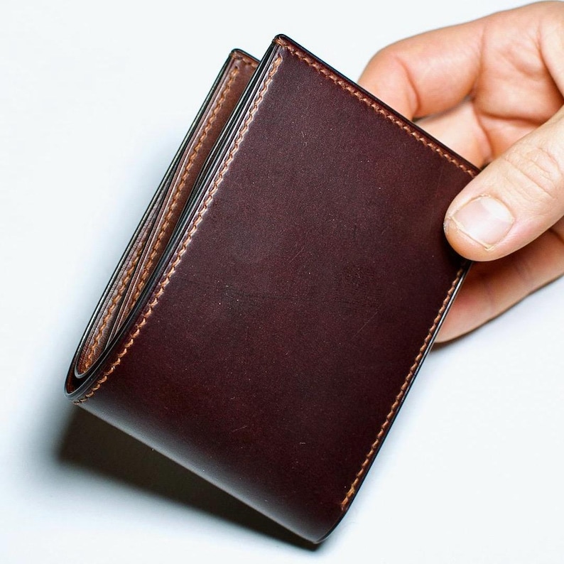 May include: A dark brown leather wallet with a bi-fold design. The wallet has a smooth, polished exterior and contrasting stitching around the edges. The interior is a lighter shade of brown.