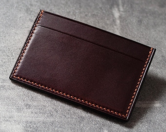 May include: A dark brown leather card holder with orange stitching. The card holder has a simple design with a single seam down the middle.