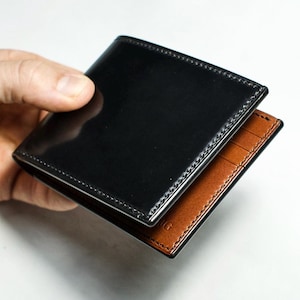 Puede incluir: Cartera bi-fold de cuero negro con acabado brillante. El interior es de color marrón en contraste con ranuras para tarjetas visibles. La cartera se sostiene abierta, mostrando su diseño. Un accesorio clásico.