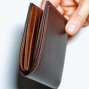 May include: A brown leather wallet with a bi-fold design. The wallet is held open, revealing multiple card slots and a lighter brown interior. The edges are stitched with a contrasting thread. The wallet appears to be new and unused.