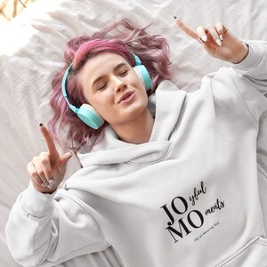 以下が含まれることがあります： 「JOMO Joy of Missing Out」と「Joyful Moments」の文字がプリントされた白いパーカー。ピンクの髪の女性がパーカーを着てヘッドホンで音楽を聴いています。