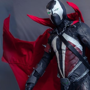 Pode incluir: Um traje de corpo inteiro do personagem Spawn com um fato texturizado em preto e vermelho escuro, um emblema branco no peito, uma capa vermelha fluida, ombreiras e braçadeiras prateadas com espinhos, uma corrente de metal e uma fivela de cinto em forma de caveira.