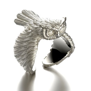 May include: A silver ring with an owl design. The owl's wings are spread out and its eyes are open. The ring is made of metal and has a smooth finish.