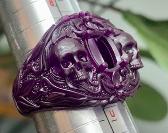 May include: A purple wax ring with a skull design and a large oval gemstone setting. The ring is on a metal ring sizer.