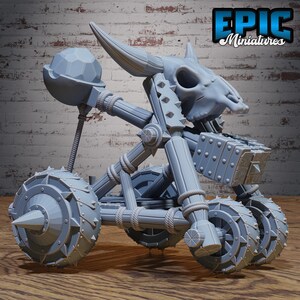 May include: A grey miniature trebuchet model featuring a skull and horns, with the text "EPIC Miniatures". The model has wheels, a launching arm, and a bucket. The design includes rope detailing and spiked accents, suggesting a fantasy or historical theme.