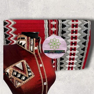 May include: A red and white patterned saddle blanket with black and silver accents. The blanket is folded in half and has a logo that says "The Aztec Couture, Ride in What You Believe". A red shirt with a decorative collar and embellishments is in the foreground.