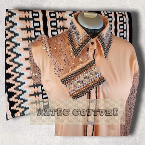 Puede incluir: Camisa de color melocotón con un diseño de lentejuelas y abalorios negros. La camisa tiene un cuello puntiagudo y cierre de botones. El texto "AZTEC COUTURE" es visible en la camisa.