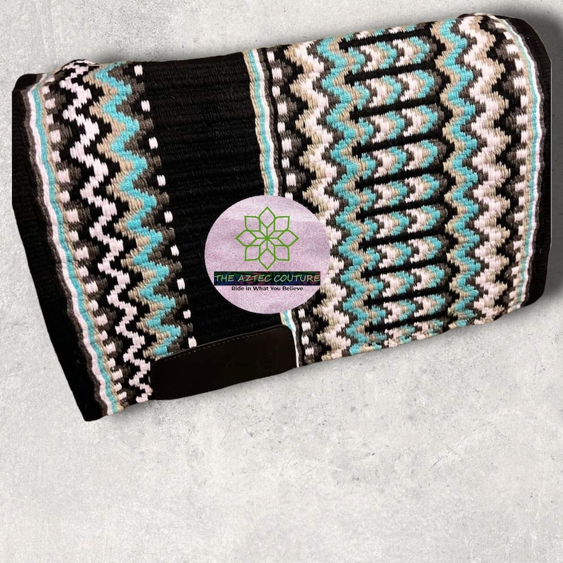 May include: Black and white saddle pad with turquoise and brown accents. The saddle pad has a geometric pattern and the text "THE AZTEC COUTURE Ride in What You Believe" is printed on the pad.