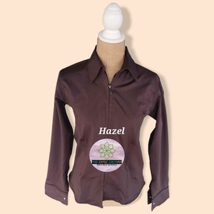 May include: A brown long-sleeve shirt with a zipper closure and a small logo on the front that says "The Aztec Couture" and "Pride in what you believe". The shirt is on a white mannequin. The word "Hazel" is written in white text above the logo.