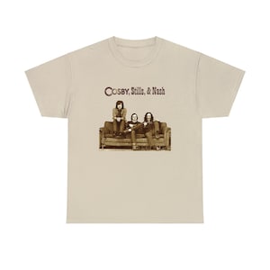 Könnte beinhalten: Ein beiges T-Shirt mit einem Vintage-Bild von Crosby, Stills & Nash, die auf einer Couch sitzen. Die Bandmitglieder tragen Freizeitkleidung und haben langes Haar. Das Bild ist in Braun- und Weißtönen gehalten.