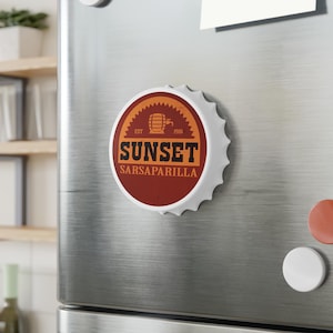 Puede incluir: Un imán de tapón de botella blanco con un diseño rojo y naranja. El diseño presenta el texto "SUNSET SARSAPARILLA" con una ilustración de barril. El imán está adherido a un refrigerador de acero inoxidable.