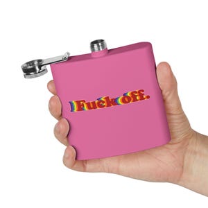 Pode incluir: Uma garrafa rosa com tampa e dobradiça prateadas. A garrafa tem o texto "Fuck off." em cores do arco-íris. A garrafa é segurada em uma mão.