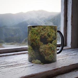 Puede incluir: Una taza de café de cerámica con una imagen de primer plano de cogollos de cannabis verdes y marrones. Sale vapor de la taza, lo que sugiere una bebida caliente. La taza está sobre un alféizar de madera pintado de blanco y desgastado, con una vista de la montaña.