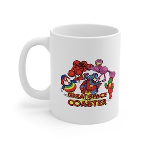 Puede incluir: Taza de cerámica blanca con una ilustración colorida de personajes de la caricatura "The Great Space Coaster". Los personajes son un perro rojo, un pájaro rosa, un elefante azul, un payaso arcoíris y un lagarto verde. El texto "The Great Space Coaster" está impreso en rojo y negro.