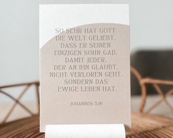 May include: A beige card with a rounded top corner sits on a woven wicker surface. The card features a quote in German, "So sehr hat Gott die Welt geliebt. Dass er seinen einzigen Sohn gab. Damit jeder, der an ihn glaubt, nicht verloren geht, sondern das ewige Leben hat." The quote is attributed to Johannes 3:16.