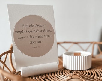 Op de afbeelding: Een witte kaart met een bruine cirkel met de tekst "Von allen Seiten umgibst du mich und hälst deine schützende Hand über mir. PSALM 139.5" op een gevlochten rieten dienblad met een witte kaars.