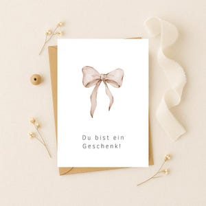 Könnte beinhalten: Eine weiße Karte mit einer hellbraunen Aquarell Schleife und dem Text "Du bist ein Geschenk!"