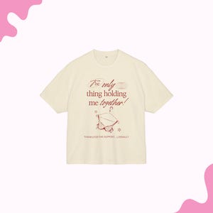 May include: Cream-colored t-shirt with the text "The only thing holding me together!" in red script. A red illustration of a bra on a hanger is below the text, with the words "THANKS FOR THE SUPPORT... LITERALLY."