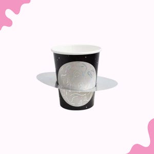 May include: A black paper cup with a white rim and a planet design. The planet is gray with silver accents. The cup sits on a silver, circular base. The background is white with pink accents.