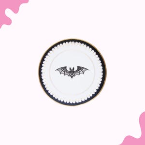 May include: White plate with a black bat design in the center. The plate has a black and gold rim with a scalloped edge. The bat is a detailed, lacy design.