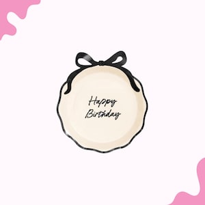 May include: A cream-colored plate with a scalloped edge and a black bow design. The words "Happy Birthday" are written in black script in the center of the plate. The plate is a party supply.