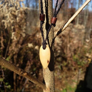 May include: A wooden pendant shaped like a teardrop hangs from a black cord necklace with brown wooden beads. The pendant is light brown with a darker brown grain pattern.