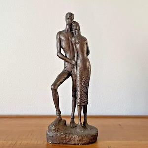 May include: A bronze sculpture of a couple embracing. The man is standing with his arm around the woman, who is leaning against him. The sculpture is on a base that resembles a rocky outcropping.