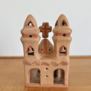 May include: A tan-colored ceramic church figurine with two bell towers and a cross on top. The towers have triangular cutouts and the structure has decorative red-brown accents. The church has two arched openings and a small bell inside each tower.