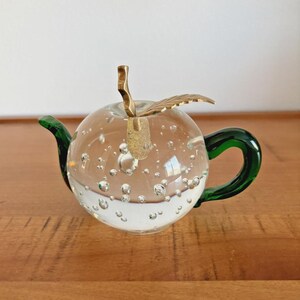 May include: A clear glass teapot with a bubble design, green handle and spout, and a gold-colored leaf and stem. The teapot is sitting on a wooden surface.