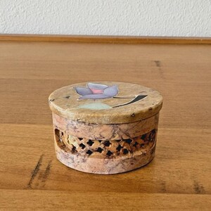 May include: A round, decorative box with a lid featuring a floral design in shades of purple, pink, and green. The box has a carved, openwork design around the sides and is made of a stone-like material.