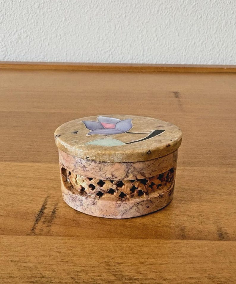 May include: A round, decorative box with a lid featuring a floral design in shades of purple, pink, and green. The box has a carved, openwork design around the sides and is made of a stone-like material.