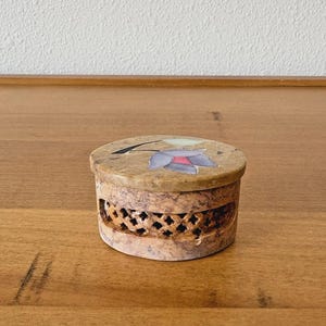 May include: A small, round, beige stone box with a floral design on the lid. The lid features a purple and red flower. The sides of the box have a decorative, openwork pattern.