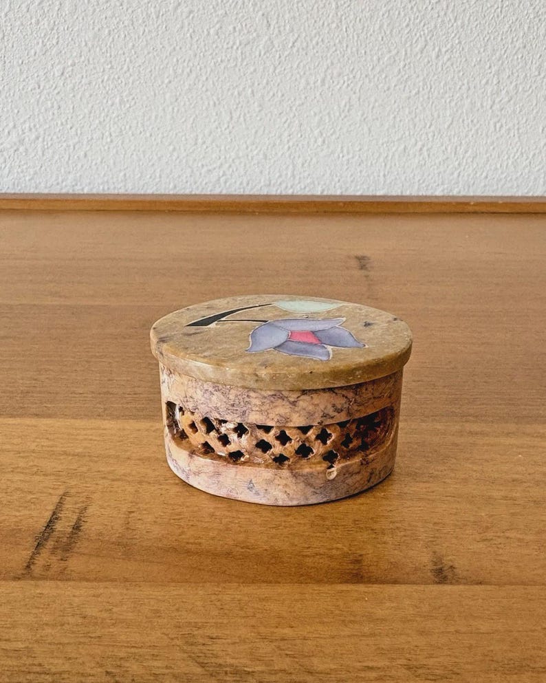 May include: A small, round, beige stone box with a floral design on the lid. The lid features a purple and red flower. The sides of the box have a decorative, openwork pattern.