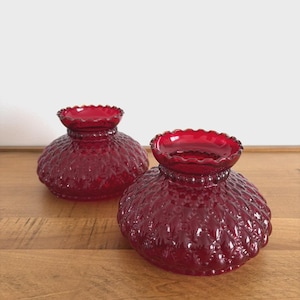 May include: Two ruby red glass vases with a textured, patterned surface and ruffled rims. The vases have a bulbous shape and sit on a wooden surface. The vases are decorative and add a touch of elegance.