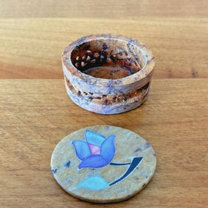 May include: A small, round, beige stone box with a matching lid. The lid features a blue flower design with a black stem. The box has a carved design around the sides. The box and lid are on a wooden surface.